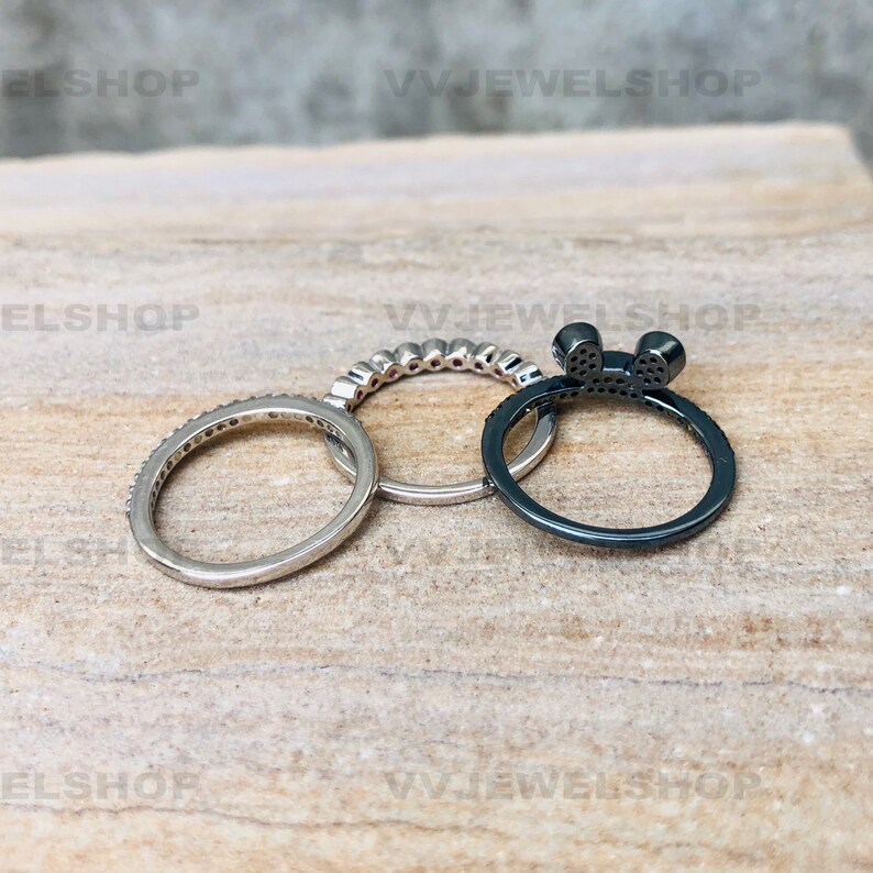 May include: Three rings on a light brown surface. One ring is silver with small stones, another is silver with a row of stones, and the third is black with a bow design and small stones. The rings are for jewelry.