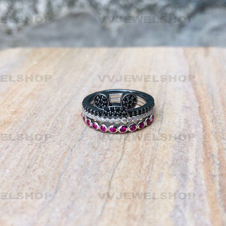 May include: Three stacked rings. The top ring is black with a Mickey Mouse head design made of black stones. The middle ring is silver with white stones. The bottom ring is silver with red stones. The rings are on a light brown surface.