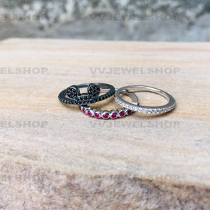May include: Three rings are displayed. One ring is black with a Mickey Mouse design, another is silver with small red stones, and the third is silver with small clear stones. The rings are on a light brown surface.