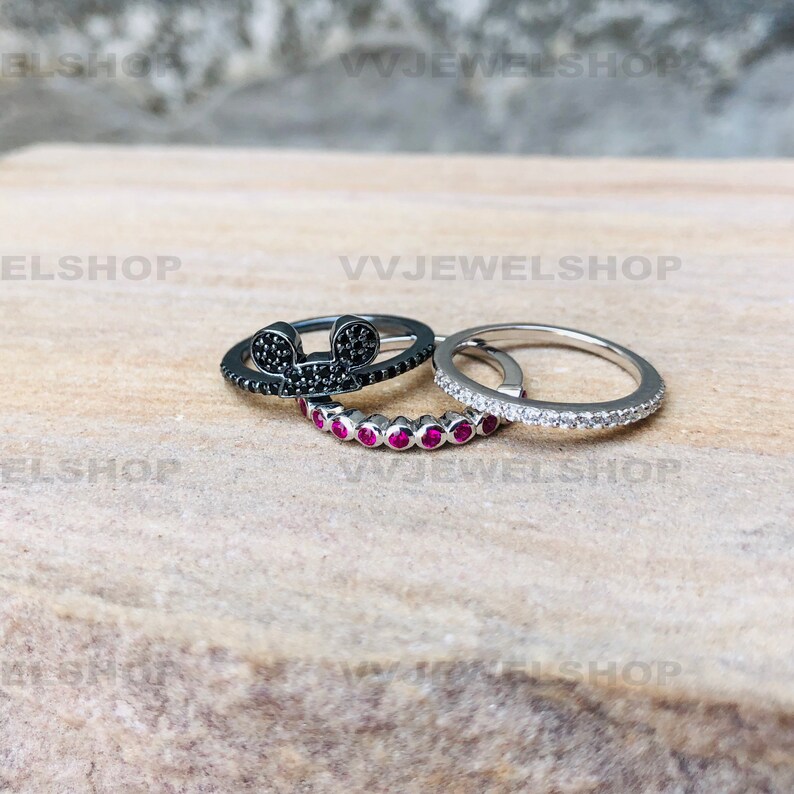 May include: Three rings are displayed. One ring is black with a Mickey Mouse design, another is silver with small red stones, and the third is silver with small clear stones. The rings are on a light brown surface.