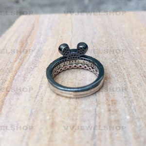 May include: A three-ring set featuring a black Mickey Mouse-shaped top ring with small black stones. The middle ring has small red stones, and the bottom ring is silver. The rings are on a light brown surface.