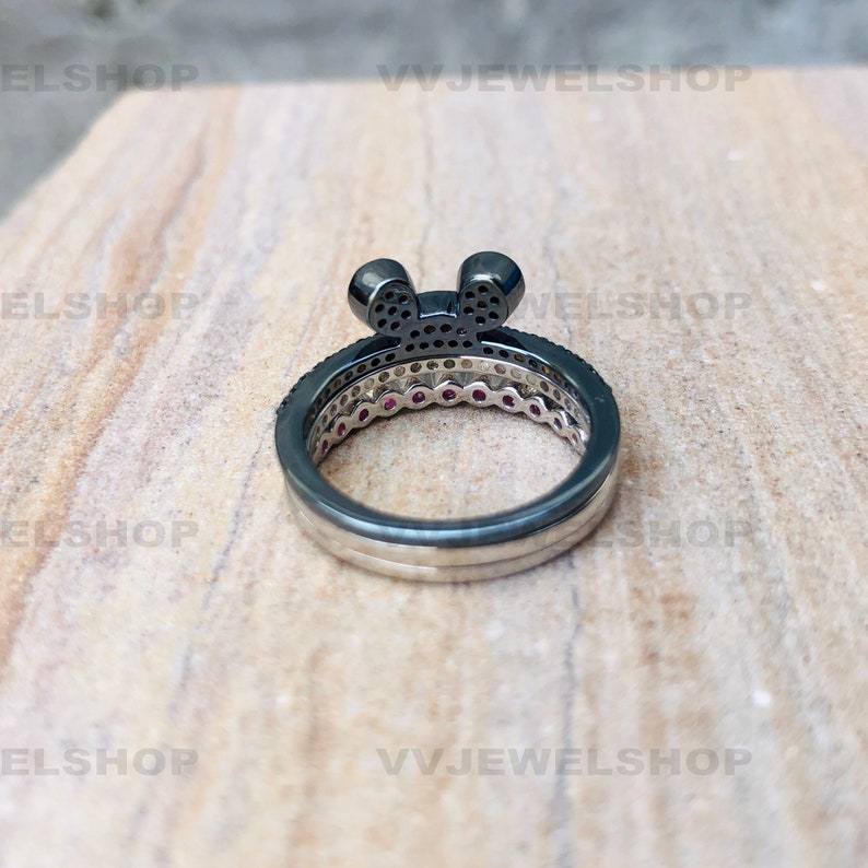May include: A three-ring set featuring a black Mickey Mouse-shaped top ring with small black stones. The middle ring has small red stones, and the bottom ring is silver. The rings are on a light brown surface.