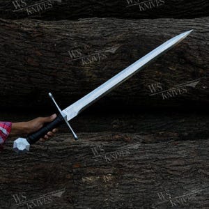 May include: A long, double-edged steel sword with a straight crossguard and a black, textured grip. The pommel is a silver-coloured, multi-faceted geometric shape. The blade is pointed and metallic, reflecting light against a dark, textured background.
