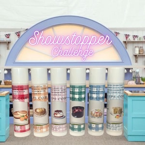 May include: Six white tubes of lip balm with different cake designs on the labels. The tubes are labeled with the text "Lips.com" and "Lip Balm LLC". The tubes are arranged in a row on a counter with a sign above them that reads "Showstopper Challenge".