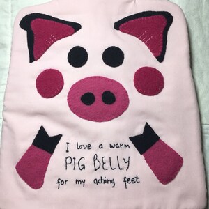 Puede incluir: Botella de agua caliente de tela rosa en forma de cerdo con ojos negros y una nariz rosa. El texto dice "Me encanta un PIG BELLY caliente para mis pies doloridos".