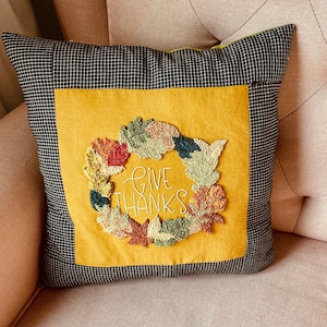 Puede incluir: Una almohada amarilla con un borde a cuadros blanco y negro. La almohada tiene un centro amarillo con una corona de hojas de colores y el texto "Give Thanks" bordado en blanco.