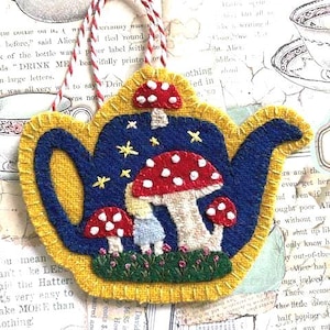 May include: A whimsical, felt teapot ornament with a blue body, yellow trim, and a red and white mushroom design. The ornament is decorated with stars and a small figure. It hangs from a red and white twine.