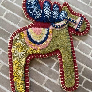 Puede incluir: Adorno artesanal de fieltro con forma de caballo Dala. El caballo es verde con ribetes rojos y está bordado con diseños florales y de hojas blancos, amarillos, rosas y azules. Se adjunta un cordón trenzado azul y blanco para colgarlo.