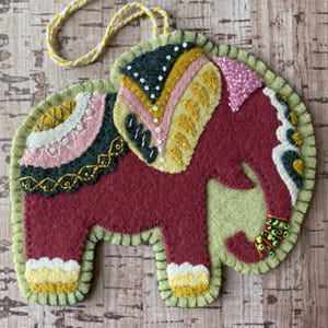 May include: A handcrafted felt elephant ornament in burgundy, green, yellow, and pink. The elephant features intricate embroidery and beadwork details, with a yellow and white cord for hanging. The ornament has a whimsical, folk art style.
