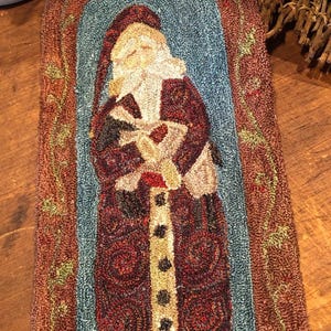 May include: A rectangular rug featuring a hooked yarn design of a figure with a white beard wearing a long dark red coat with black buttons. The figure is centered against a light blue background, framed by a textured brown border with green vines.