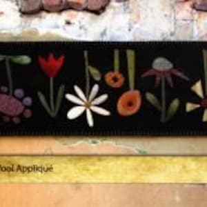 May include: A black wool applique banner featuring a garden row design with various colorful flowers. The banner is 11 inches by 60.5 inches. The design includes a daisy, tulip, and other floral elements. The banner is labeled "211 - Garden Row".