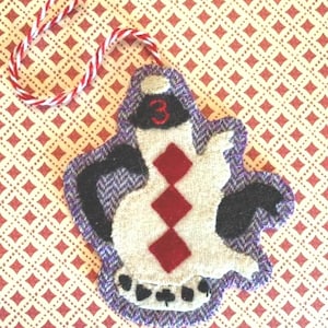 Könnte beinhalten: Handgefertigter Filzschmuck in Form einer Spielkarte mit weißer Mitte und drei roten Rauten. Der Schmuck hat einen lila Tweed-Rand und eine rot-weiße Schnurschlaufe zum Aufhängen.