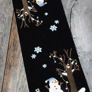 May include: A black table runner featuring two white snowmen, each wearing a blue hat and standing near a brown tree decorated with colorful string lights. The design includes white snowflakes, a small shovel, and a red sled on the snow.