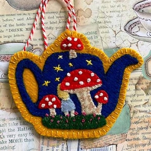 May include: A whimsical, felt teapot-shaped ornament with a red and white striped hanging cord. The blue teapot design features mushrooms, stars, and a small figure, all outlined in yellow.