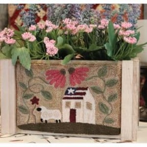 May include: A wooden planter box with a floral arrangement. The box features a stitched design of a house with an American flag, a sheep, and flowers. The arrangement includes pink and purple flowers.