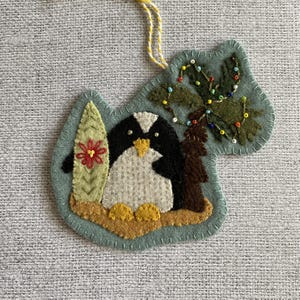 Może przedstawiać: Filcowa ozdoba świąteczna przedstawiająca pingwina z deską surfingową i ozdobionym drzewkiem. Pingwin jest czarno-biały z żółtymi stopami i dziobem. Deska surfingowa jest zielona z czerwonym kwiatem. Drzewko jest brązowe z kolorowymi koralikami.