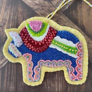 May include: A handcrafted, felt elephant ornament with intricate embroidery. The elephant is primarily blue with colorful accents of pink, green, red, and white. It has a yellow border and a yellow and white string for hanging. The ornament is a unique, decorative item.