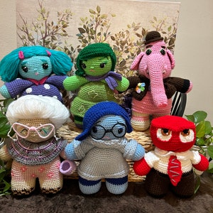 May include: Six crocheted stuffed toys representing the emotions from the movie Inside Out. The toys are sitting on a brown fuzzy surface. The characters are Joy, Sadness, Anger, Disgust, Fear, and Bing Bong.
