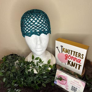 May include: A teal crocheted hat is displayed on a white mannequin head. The hat has a scalloped edge and an open, net-like design. A sign that says "Knitters gonna Knit" is visible, along with a sign that says "100% Handmade with Love".