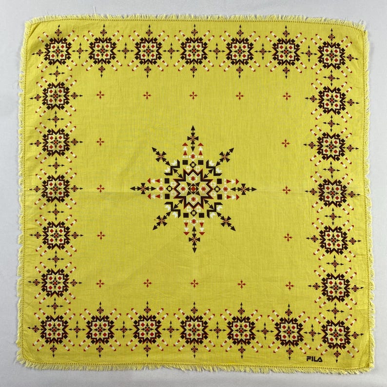 May include: A square, yellow handkerchief with a geometric pattern. The design features a central starburst and a border of repeating floral motifs in black, white, and red. The edges are fringed, and the word "FILA" is printed in the bottom right corner.