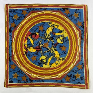 May include: A blue bandana with a colorful, circular design featuring multiple figures and a repeating pattern of stars and concentric circles. The bandana has a red, yellow, and brown border.