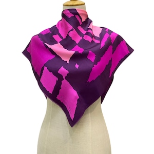 May include: A square silk scarf featuring a bold geometric pattern in shades of deep purple, vibrant magenta, and light pink. The fabric displays a repeating abstract block design with the text Christian Dior printed in the lower corner area.