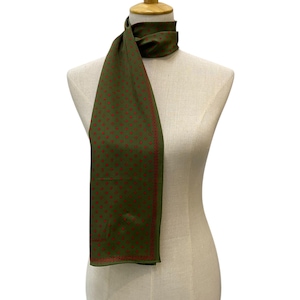 Pode incluir: Lenço verde-oliva com um padrão de pontos vermelhos repetidos e uma borda vermelha. O lenço está drapeado sobre um manequim. O lenço tem formato retangular e parece ser feito de um tecido leve.