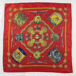 May include: A red silk scarf with a repeating pattern of drums, drumsticks, and decorative elements in gold, blue, and yellow. The scarf has a border with gold chain-like details.