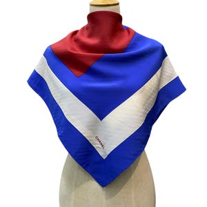 Puede incluir: Un pañuelo de seda con un diseño geométrico en rojo, blanco y azul. El pañuelo se exhibe en un maniquí. La palabra "CHANEL" está impresa en negro en el pañuelo.