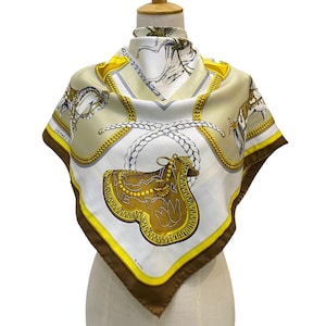 Puede incluir: Un pañuelo de seda con un diseño de silla de montar. El pañuelo presenta un fondo blanco con detalles en beige, marrón y amarillo. El diseño incluye una silla de montar, cuerda y detalles de cadena. El pañuelo se exhibe en un maniquí.