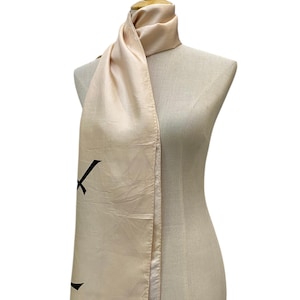 May include: A pale peach-coloured satin scarf draped over a mannequin. The scarf features a smooth, lustrous finish and a visible black graphic print on the lower section. The edges of the scarf are finished with a delicate, fine fringe detail.