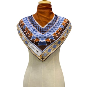 May include: A vintage Christian Dior silk scarf on a mannequin. The scarf has a geometric pattern in blue, brown, and tan. The square scarf is labelled with the Christian Dior logo. The mannequin is on a white background.