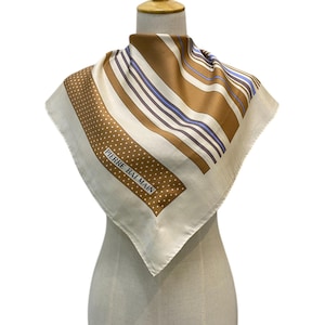 Pode incluir: Lenço quadrado de seda com fundo creme e um padrão de riscas castanhas e brancas. O lenço tem uma secção pontilhada com o texto "PIERRE BALMAIN". O lenço é exibido num manequim.