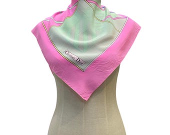 May include: A square silk scarf featuring a vibrant pink border and a central pale green panel with geometric lines. The scarf displays the text Christian Dior in a rectangular frame on the lower left side, draped over a mannequin torso.
