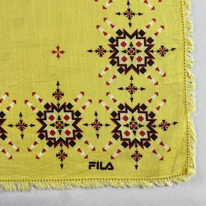 May include: Yellow square textile with a geometric pattern in black, red, and white. The design features starburst and cross-like shapes. The word "FILA" is printed in white. The edges are fringed.