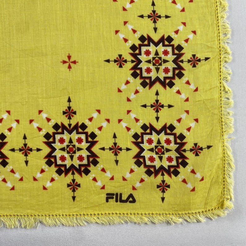 May include: Yellow square textile with a geometric pattern in black, red, and white. The design features starburst and cross-like shapes. The word "FILA" is printed in white. The edges are fringed.