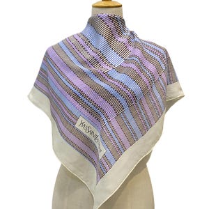 May include: A square silk scarf with a geometric pattern in shades of lavender, blue, and brown. The scarf has a white border and the text "YVES SAINT LAURENT" is visible. The scarf is displayed on a mannequin.