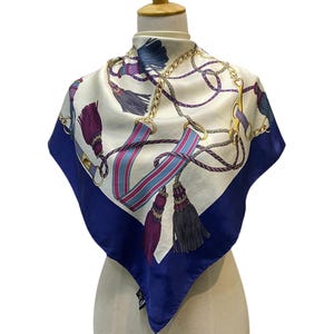 May include: Square silk scarf with a cream background, featuring a pattern of gold chains, tassels, and blue and pink striped accents. The scarf has a deep blue border and is displayed on a mannequin.