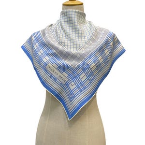 May include: A square scarf featuring a white base with a blue and grey grid pattern. The design includes a border of blue and grey lines. The text JACQUES HEIM is printed in black on the lower left side of the scarf. The scarf is draped on a mannequin.