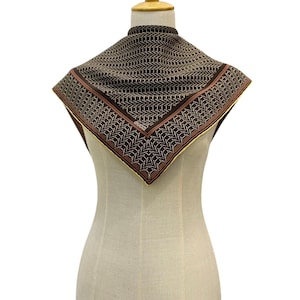 May include: A square scarf with a geometric pattern in shades of brown, beige, and red is displayed on a mannequin. The scarf has a border with a gold trim. The scarf is made of a soft, flowing fabric.