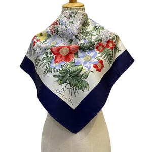 May include: A square silk scarf featuring a vibrant botanical print of red, blue, and white flowers with green foliage on a cream background. The scarf has a wide navy blue border and the text Christian Dior printed in black on the lower corner.