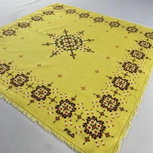 May include: A square, yellow scarf with a geometric starburst pattern in black, red, and white. The scarf has a fringed edge and the word "FILA" is printed in black in the lower right corner.