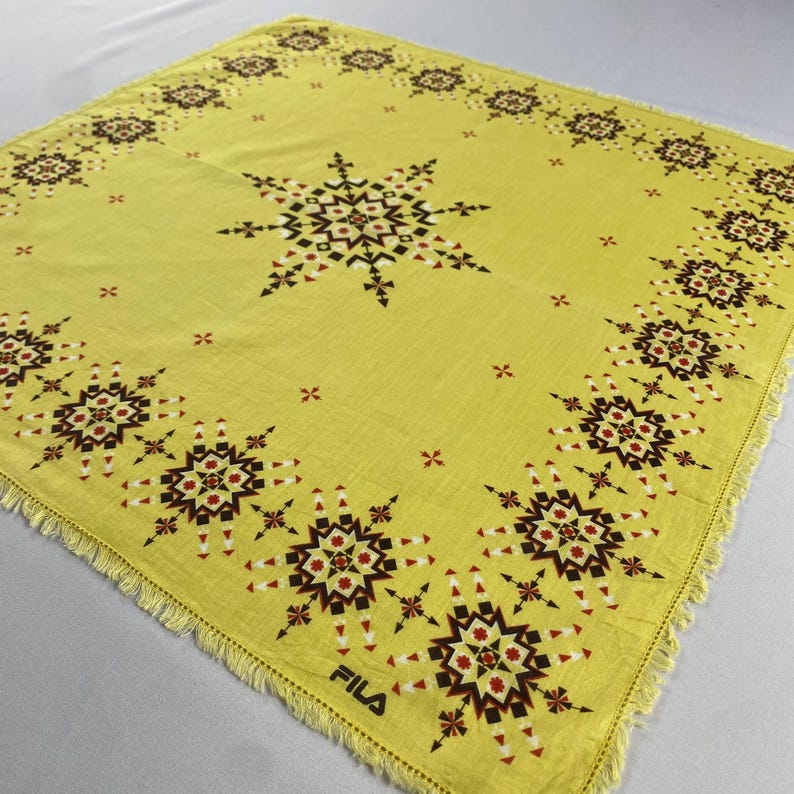 May include: A square, yellow scarf with a geometric starburst pattern in black, red, and white. The scarf has a fringed edge and the word "FILA" is printed in black in the lower right corner.