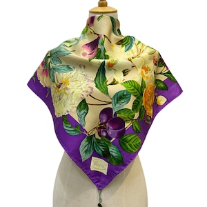 May include: A square silk scarf featuring a cream-colored center with a vibrant botanical print of white peonies, yellow roses, green leaves, and purple plums. The scarf has a wide, solid purple border and a Gucci label stitched in the corner.