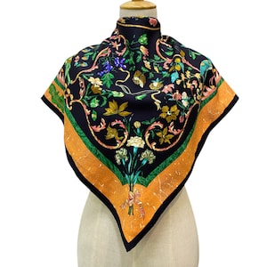 Könnte beinhalten: Ein quadratisches Seidentuch mit schwarzem Hintergrund und floralem Design. Der Schal hat einen orangefarbenen und grünen Rand mit einem Blumenstrauß und einer Schleife am unteren Rand. Der Schal wird auf einer Schaufensterpuppe präsentiert.