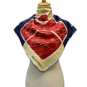 Puede incluir: Un pañuelo cuadrado de seda con un diseño geométrico audaz en rojo, blanco y azul marino. El pañuelo presenta el nombre de la marca "BALENCIAGA" en texto blanco a lo largo de un borde. El pañuelo se exhibe en un maniquí.