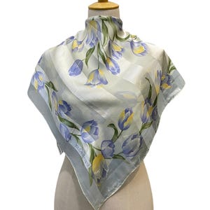 May include: A square silk scarf featuring a light green background with diagonal stripes and a floral pattern of purple and yellow tulips. The scarf displays the Yves Saint Laurent brand name printed in white text near the bottom corner.