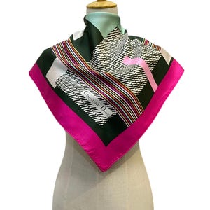 Pode incluir: Um lenço quadrado de seda com fundo verde escuro, um padrão geométrico de traços brancos, listras multicoloridas e uma borda rosa brilhante. O texto Christian Dior está impresso em branco no lenço, que está drapeado em um manequim.