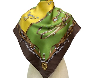 Può includere: Foulard quadrato in seta con un design equestre. La sciarpa presenta un design a blocchi di colore con tonalità gialle, verdi e marroni. La sciarpa ha un motivo di finimenti, catene e fibbie. La sciarpa è esposta su un manichino.