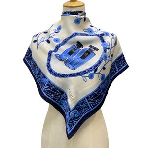 Könnte beinhalten: Ein weißer Seidenschal mit einem blauen und weißen Blumenmuster und einem blauen und goldenen Design von zwei Parfümflakons. Der Schal hat einen dunkelblauen Rand mit einem weißen "LANVIN PARIS" Logo.